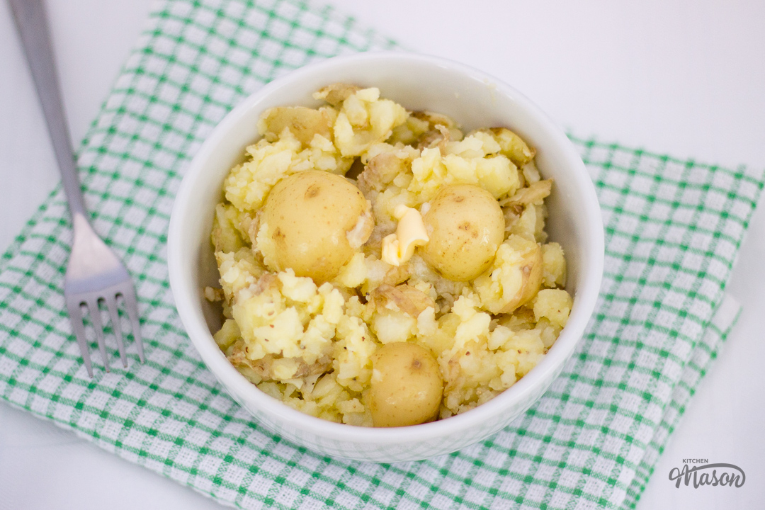 Crushed new potatoes and butter in a bowl over a green tea towel, with a fork to the side.