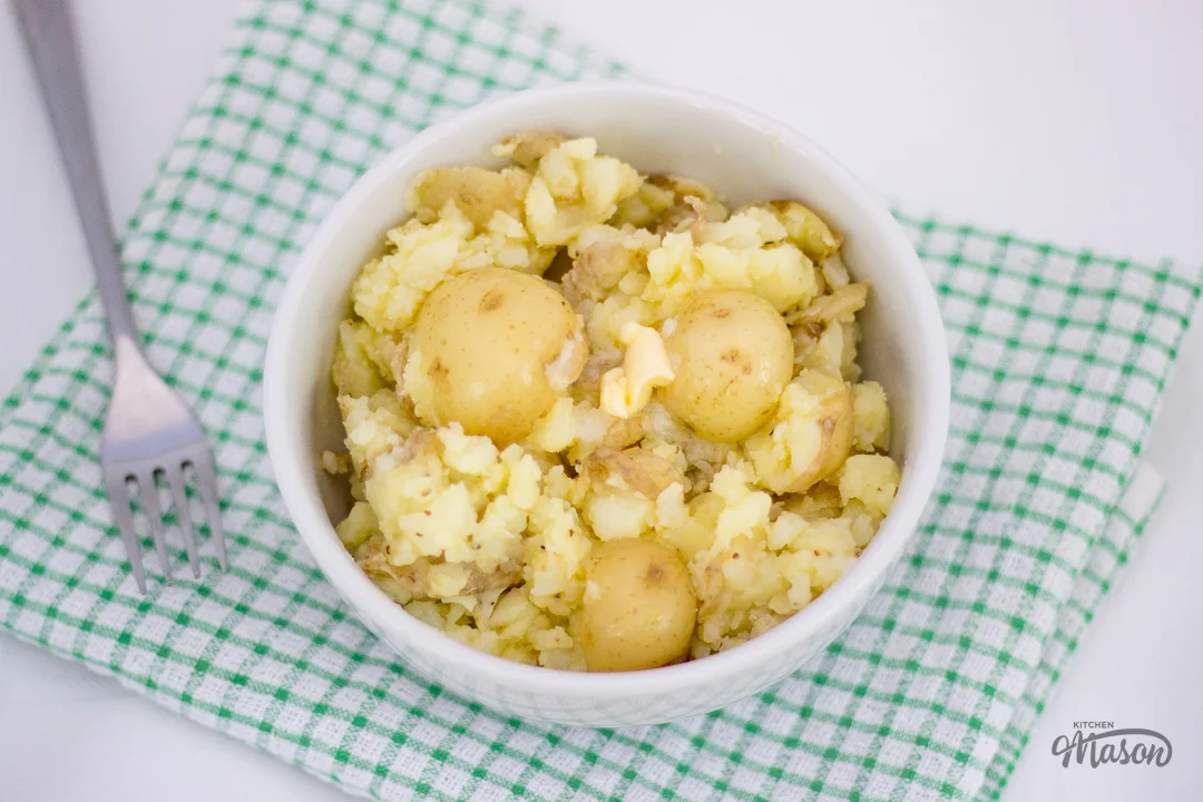 Crushed new potatoes and butter in a bowl over a green tea towel, with a fork to the side.
