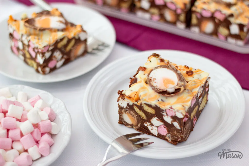 Two Creme Egg rocky road bars on white plates, with a bowl of mini marshmallows to the side.