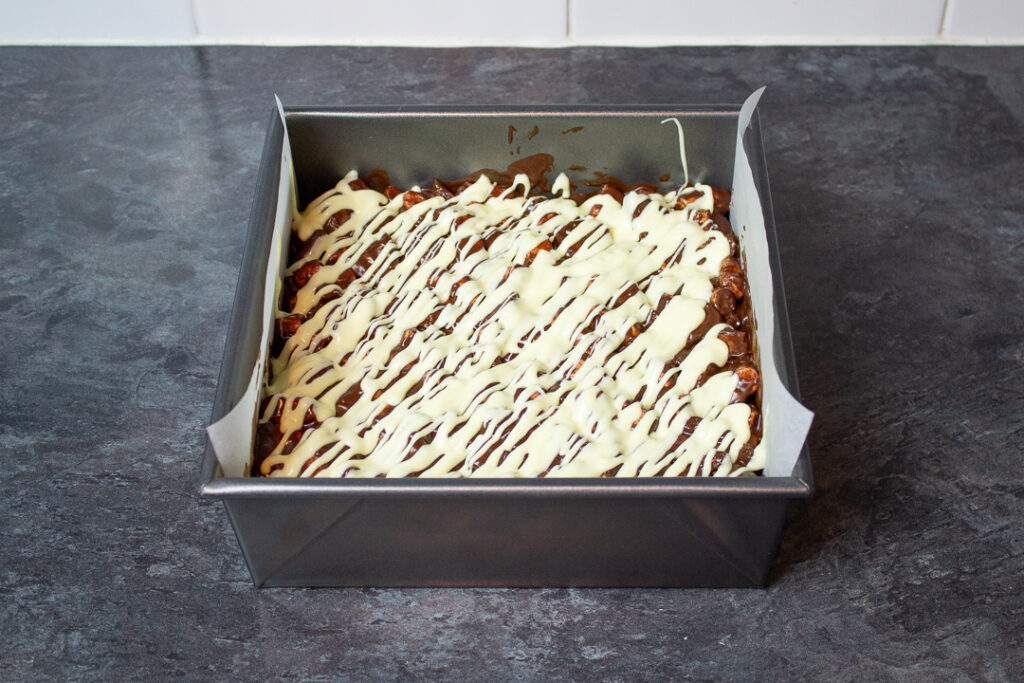 Melted white chocolate drizzled over the top of rocky road in a baking tray.