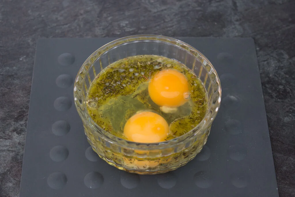 Butter, thyme, eggs, salt, and pepper in a glass ramekin on a kitchen worktop.
