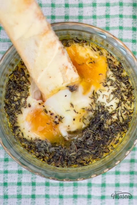 A buttered toast finger dipped into a glass ramekin filled with thyme baked eggs.