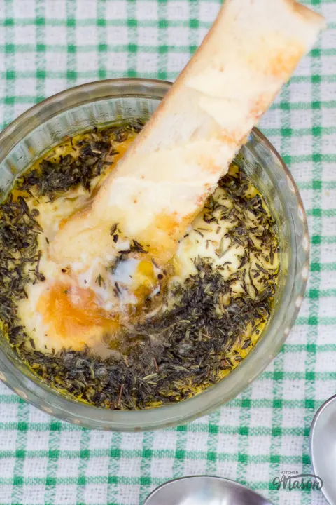 Buttered toast dipped into runny thyme baked eggs with a spoon at the side.