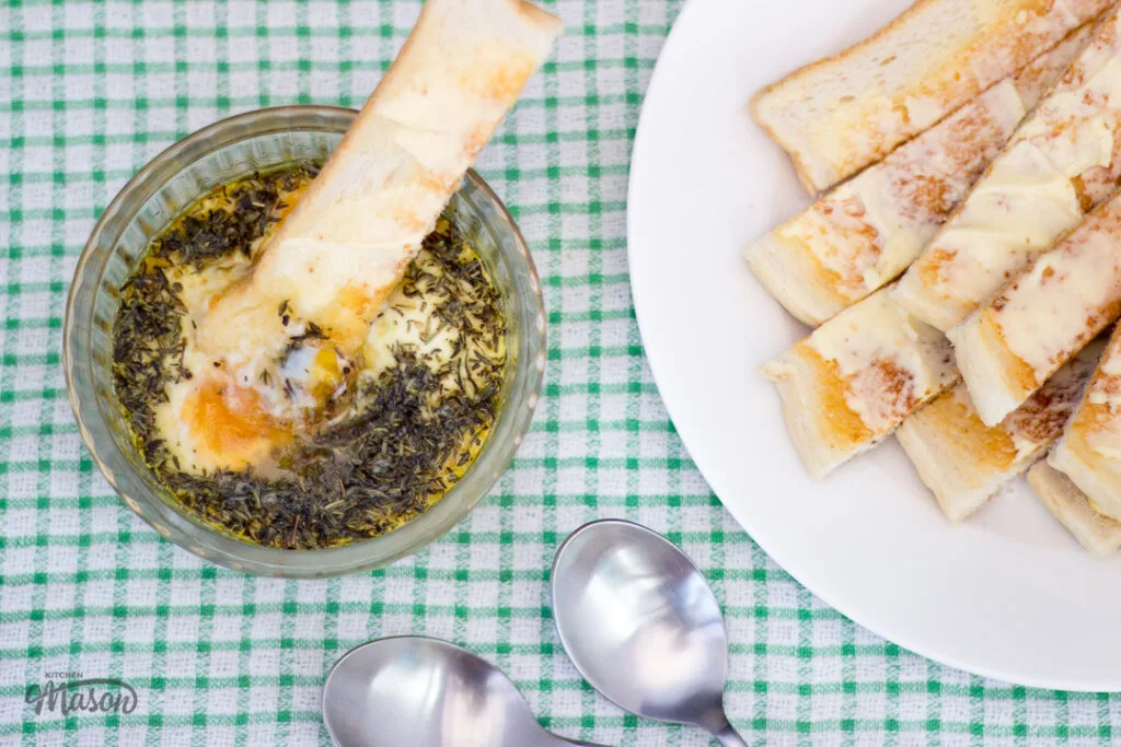 Toast dunked into thyme baked eggs in a glass ramekin, with spoons and a plate of buttered toast to the side.