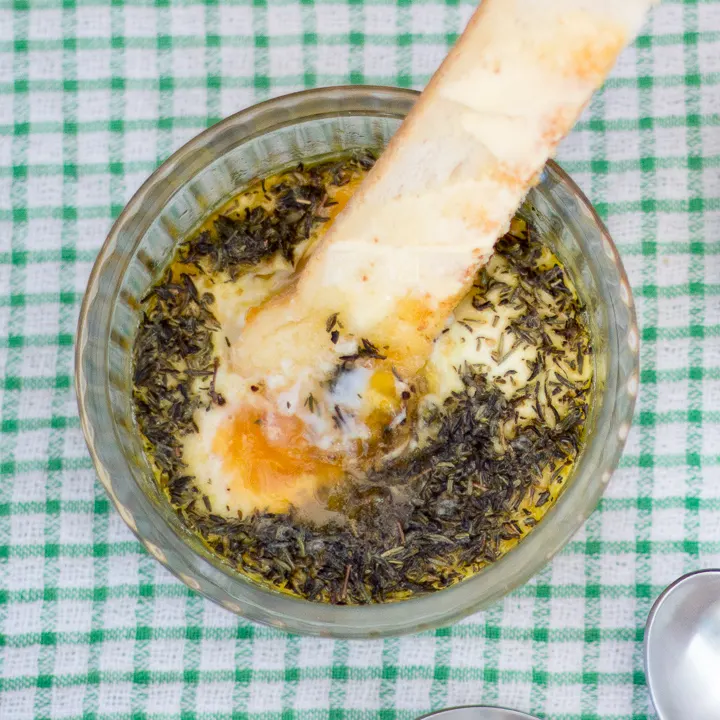 Buttered toast being dipped into thyme baked eggs in a glass ramekin.