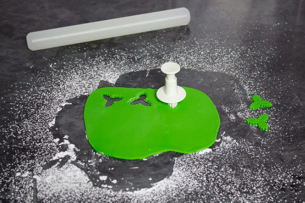Green fondant icing being shaped into holly leaves with a plunge cutter.