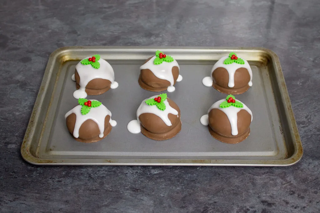 Tunnocks teacakes on a baking tray topped with runny white icing and holly and ivy sugar decorations.