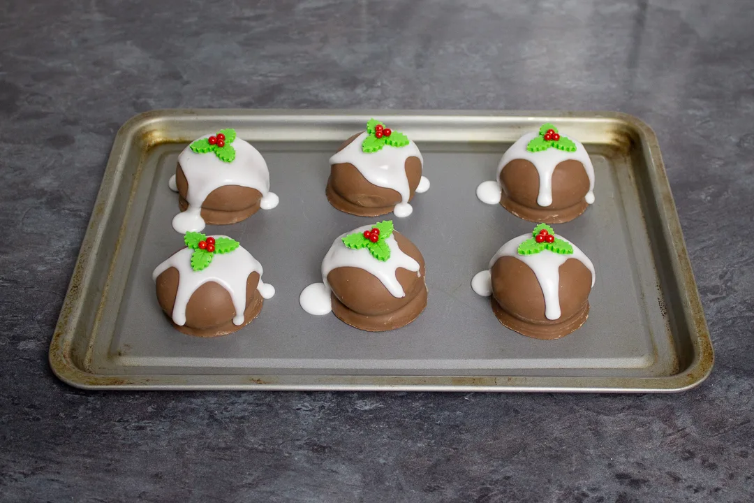 Tunnocks teacakes on a baking tray topped with runny white icing and holly and ivy sugar decorations.