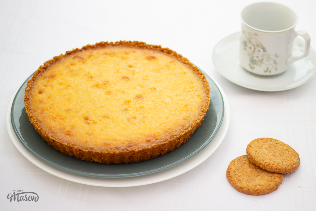 A white chocolate lemon tart on a serving plate with a cup of tea to the side.