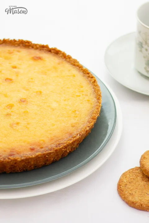A white chocolate lemon tart on a serving plate with spare biscuits and a cup of tea to the side.