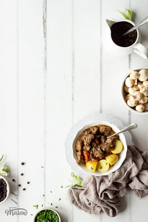 A white bowl of beef casserole and potatoes on a brown napkin. Bowls of other ingredients to the side.