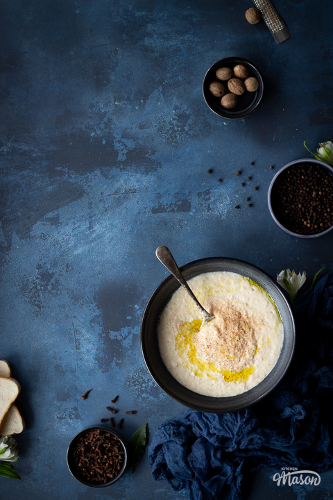 A blue bowl of bread sauce with a metal spoon in it on a blue worktop.