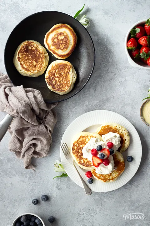 A plate of drop scones topped with berries and cream, next to a frying pan with 3 dropped scones in it.