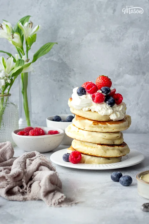 5 stacked drop scones topped with berries and cream. There are bowls of different berries to the side.