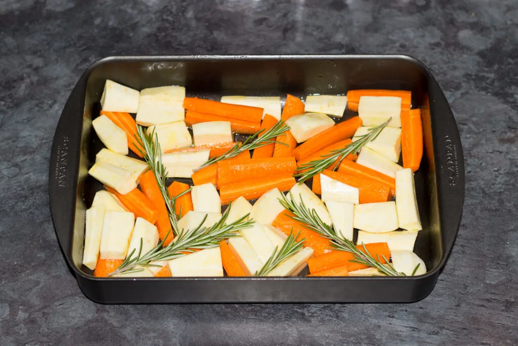 Chopped, oiled and seasoned parsnips and carrots topped with honey and rosemary in a roasting pan.