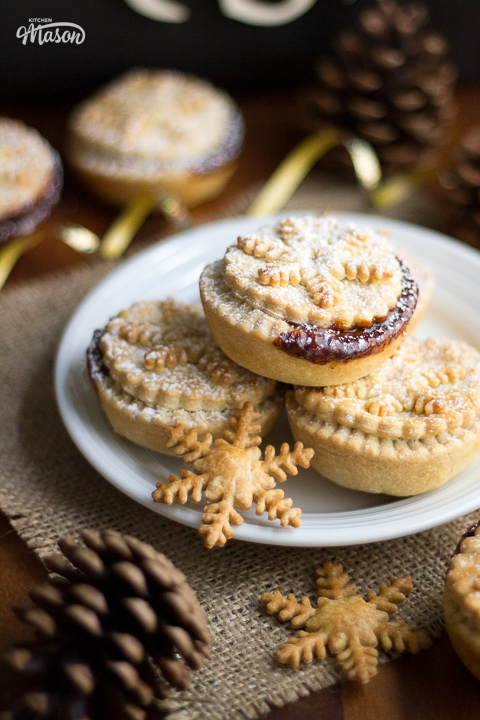 A plate topped with a few shortcrust pastry mince pies. Pine cones and gold ribbon are at either side.
