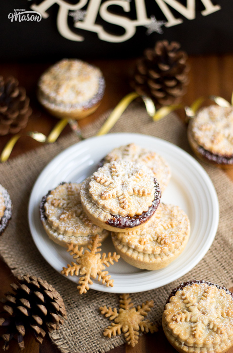 A plate of mince pies surrounded by more mince pies, pine cones, and gold curling ribbon.