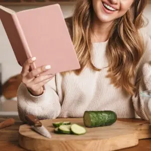 A smiling lady holding a cookbook chopping a cucumber.