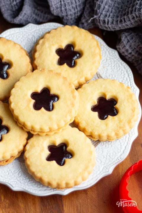 Lots of black cherry jammy dodgers on a white plate with a grey tea towel to the side.