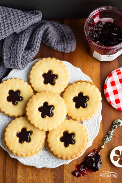 An open jar of jam next to a decorative plate filled with jammy dodgers, with a spoon of black cherry jam at the side.