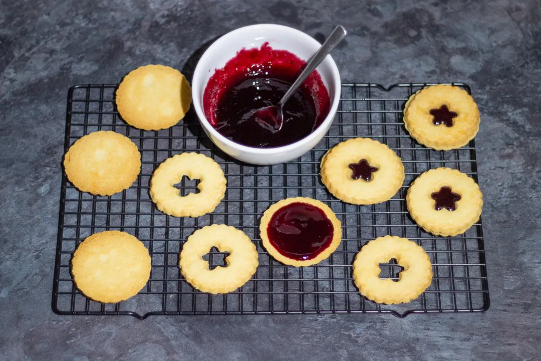 Baked biscuits being filled and sandwiched with black cherry jam on a wire cooling rack.