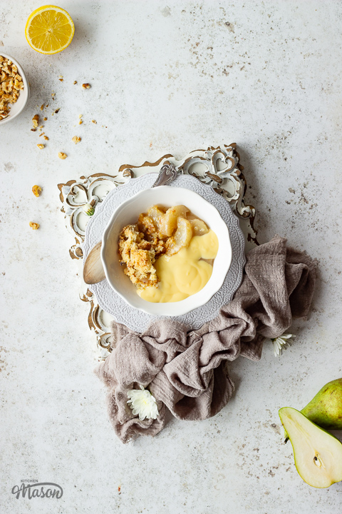 A white bowl filled with pear crumble and custard, set on a wooden board with a linen napkin at the side.