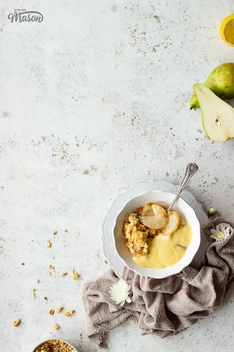 A bowl of pear crumble and custard with cut pears and lemons to the side.