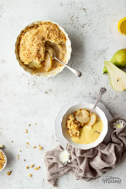 A baking dish of pear crumble with a serving spoon, next to a bowl of pear crumble and custard. There are chopped walnuts, cut pears and lemons on the worktop too.