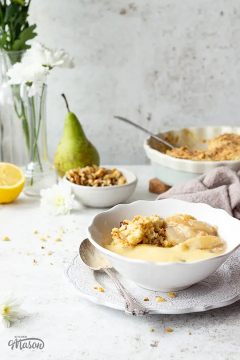 A bowl of pear crumble and custard on a plate with a spoon. There is a vase of pretty white flowers in the background.