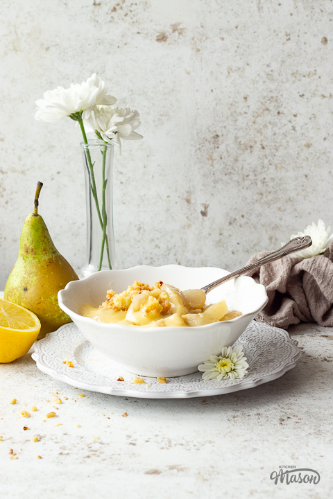 A bowl of pear crumble and custard with a spoon, set on a decorative plate.