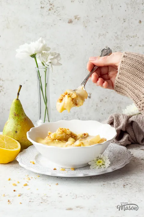 Someone spooning pear crumble and custard from a bowl. The custard is dripping and it looks very tasty!