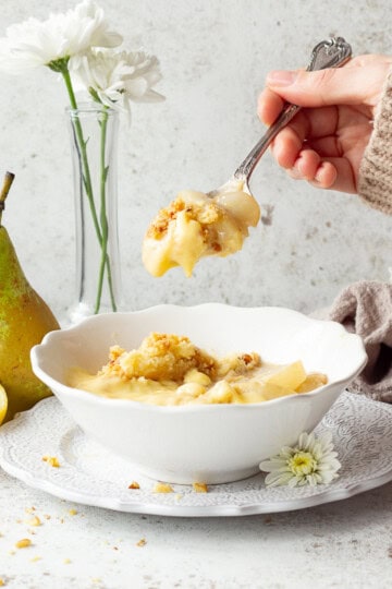 Someone holding a spoonful of pear crumble and custard above a bowl full.