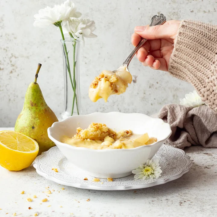 Someone holding a spoonful of pear crumble and custard above a bowl full.