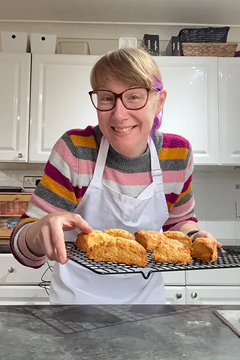Emma Mason smiling and holding a cooling rack filled with freshly baked cheese scones from one of her cook-alongs.