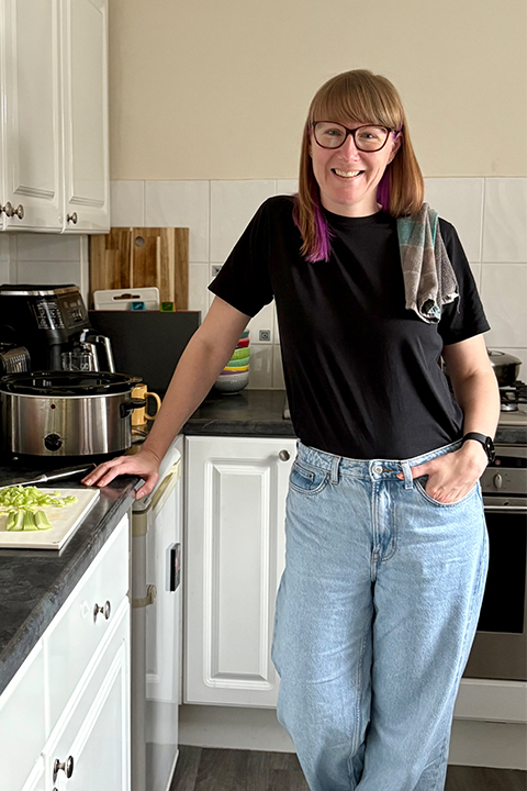 A happy Emma Mason in her home kitchen, with a chopping board filled with vegetables to the side of her on her kitchen worktop.