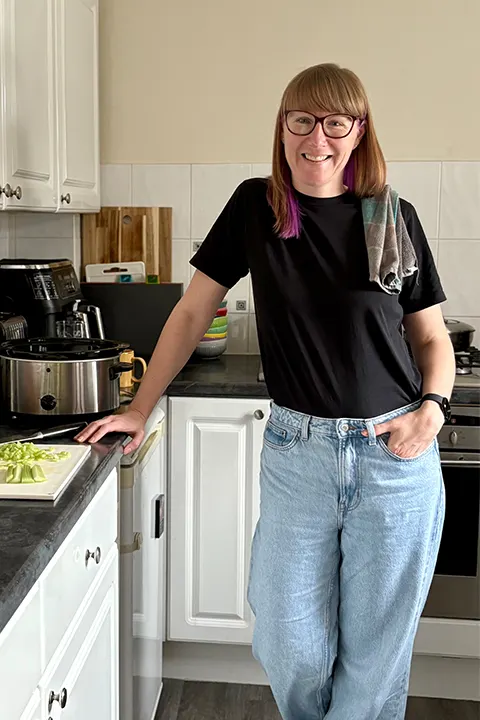 A happy Emma Mason in her home kitchen, with a chopping board filled with vegetables to the side of her on her kitchen worktop.