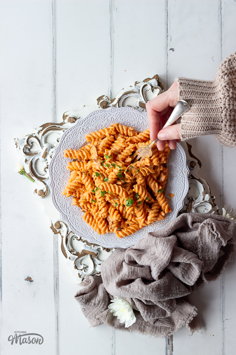 Someone pushing a fork into a plate of bacon pasta topped with chopped parsley.