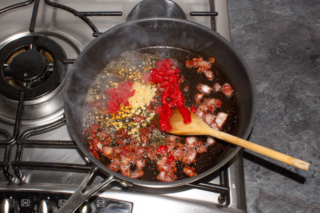 Tomato puree and garlic being cooked out in a frying pan with bacon.