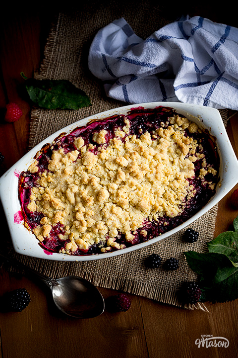 A vibrant Summer fruit crumble in a white dish surrounded by blackberries and raspberries.