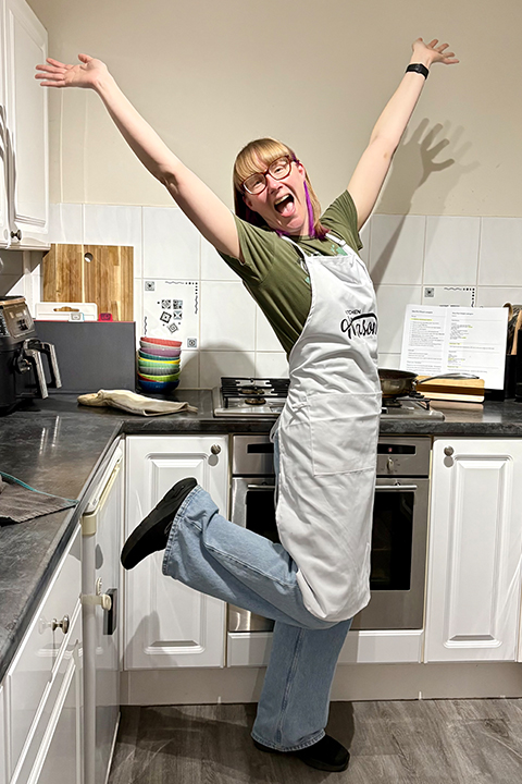 A very happy Emma raising her arms and leg in joy, in her kitchen wearing a Kitchen Mason apron.