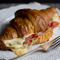 A warm and melted gouda and prosciutto croissant on a white plate with a fork.