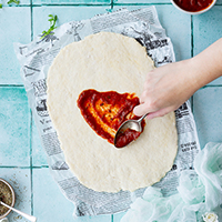 Quick pizza dough rolled out on a worktop being spread with a tomato sauce.