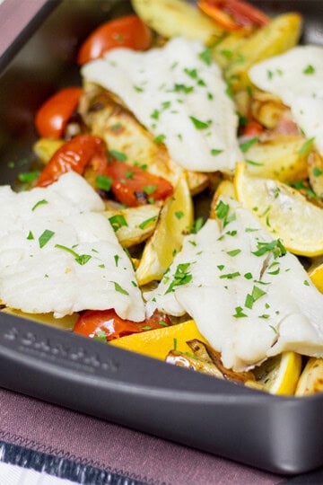 Oven baked fish, chips, tomatoes and lemon in a large roasting dish.