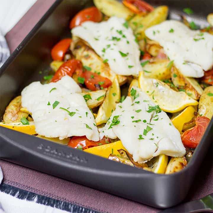 Oven baked fish, chips, tomatoes and lemon in a large roasting dish.