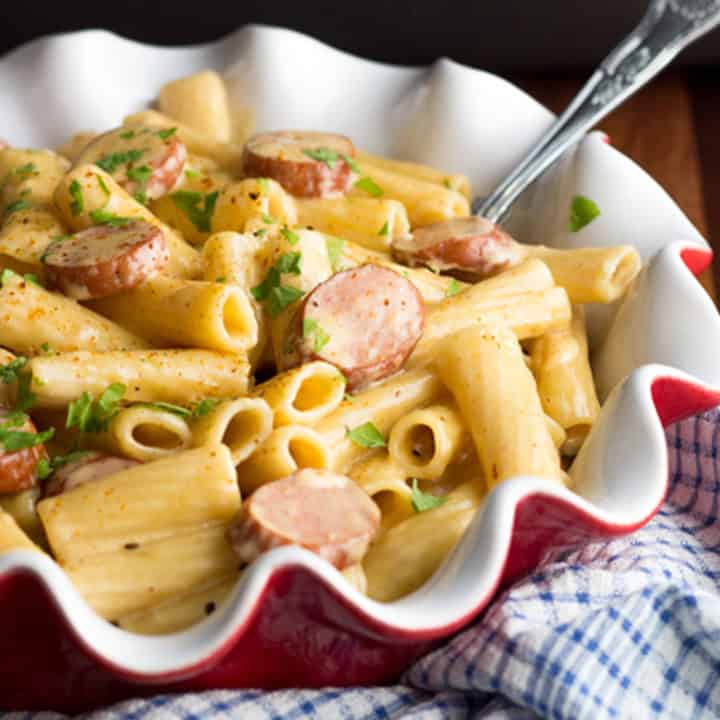 A fluted red and white dish filled with cajun sausage pasta. A large serving spoon is sticking out the side.