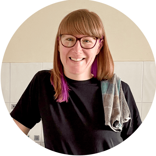 Emma smiling standing in her kitchen with a black t-shirt on and a tea towel slung over her shoulder.